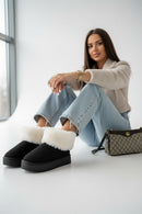 Mujer sentada vistiendo botín borreguita negro con pantalones de mezclilla y bolso elegante, mostrando estilo y comodidad.